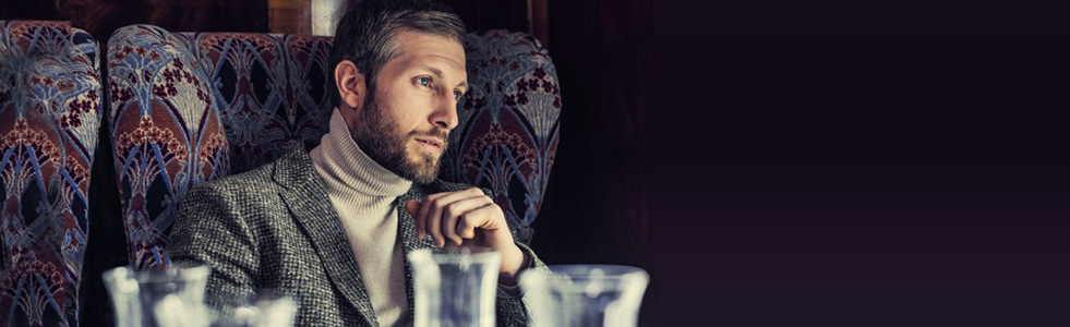 Ein Man sitzt nachdenklich in einem Sessel. Er trägt einen Rollkragenpullover und ein Sakko.