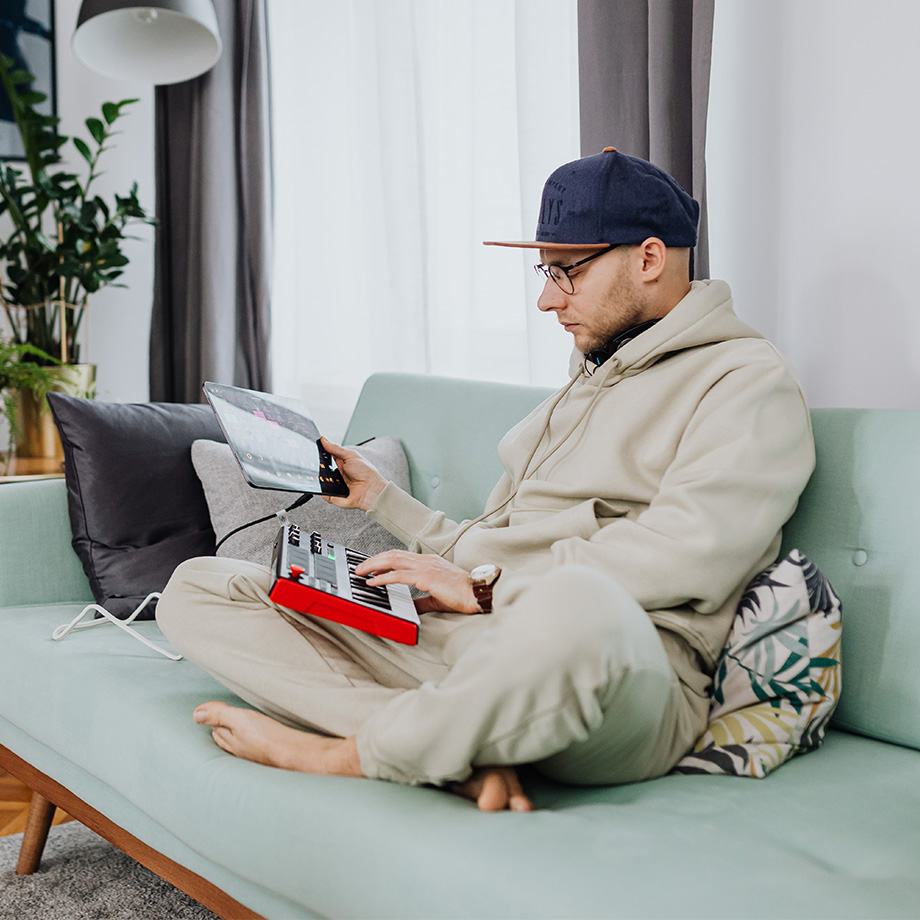 Das Bild zeigt einen Mann in einem beigefarbenen Hoodie und einer Baseballkappe, der auf einem Sofa sitzt, ein Tablet in der Hand hält und ein Musik- oder DJ-Controller-Gerät auf seinem Schoß bedient.