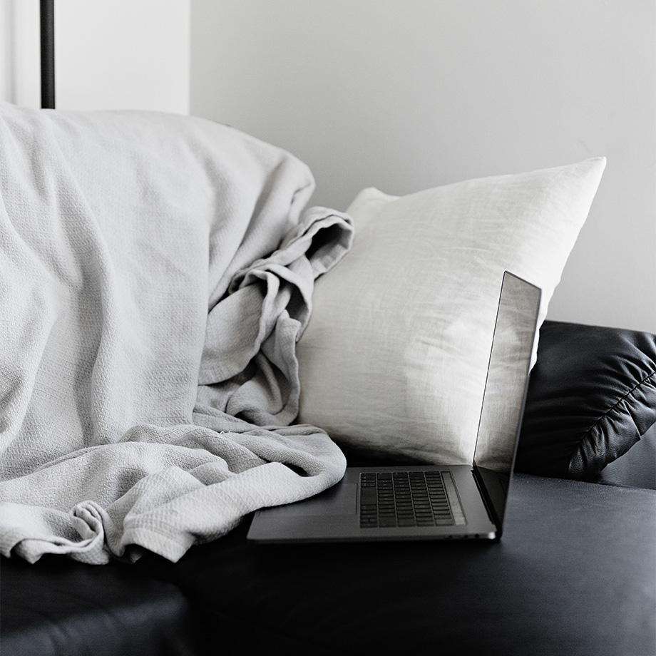 Das Bild zeigt einen Laptop auf einem schwarzen Sofa, neben einem weißen Kissen und einer hellen Decke.