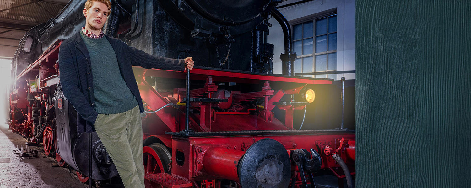 Ein Mann lehnt lässig an einer schwarz-roten Dampflokomotive. Er trägt einen dunkelblauen Cardigan, darunter einen grünen Pullover, ein rot kariertes Hemd und eine grüne Cordhose. Rechts ist ein grüner Cordstoff sichtbar.