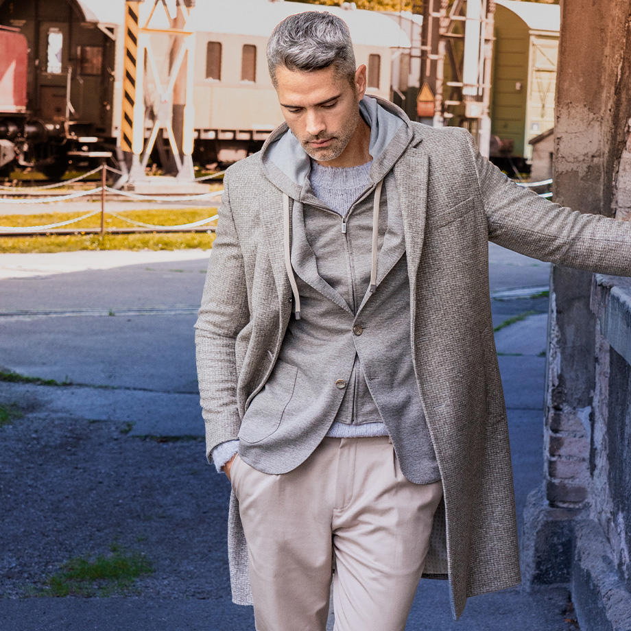 Ein männliches Model zeigt einen modernen Layering-Look, der aus einem grauen Strickpullover, einem grauen Sakko mit Kapuze und einem beige-grauen Mantel besteht, waehrend das Model mit einer Hand laessig an einer Wand lehnt und in einer urbanen, aeusseren Umgebung posiert.