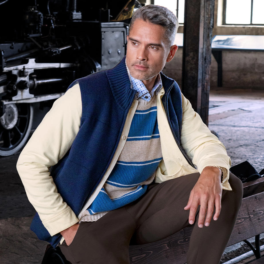 Ein männliches Model steht im Bahnpark und praesentiert ein stilvolles Layering-Outfit, das aus einem gemusterten Hemd, einem blau-beige gestreiften Pullover, einer blauen Weste und einem beigen Overshirt besteht, das mit einer braunen Hose kombiniert ist, wodurch der Look eine harmonische Farbpalette und laessige Eleganz ausstrahlt.