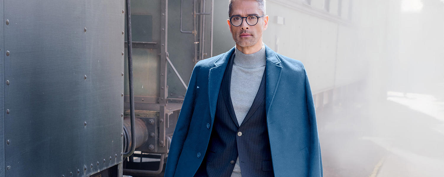 Ein Mann mit grauem Haar und Brille trägt einen eleganten, dunkelblauen Mantel über einem dunkelblauen Anzug und einem grauen Rollkragenpullover. Er steht neben einem Zug in nebliger Umgebung.
