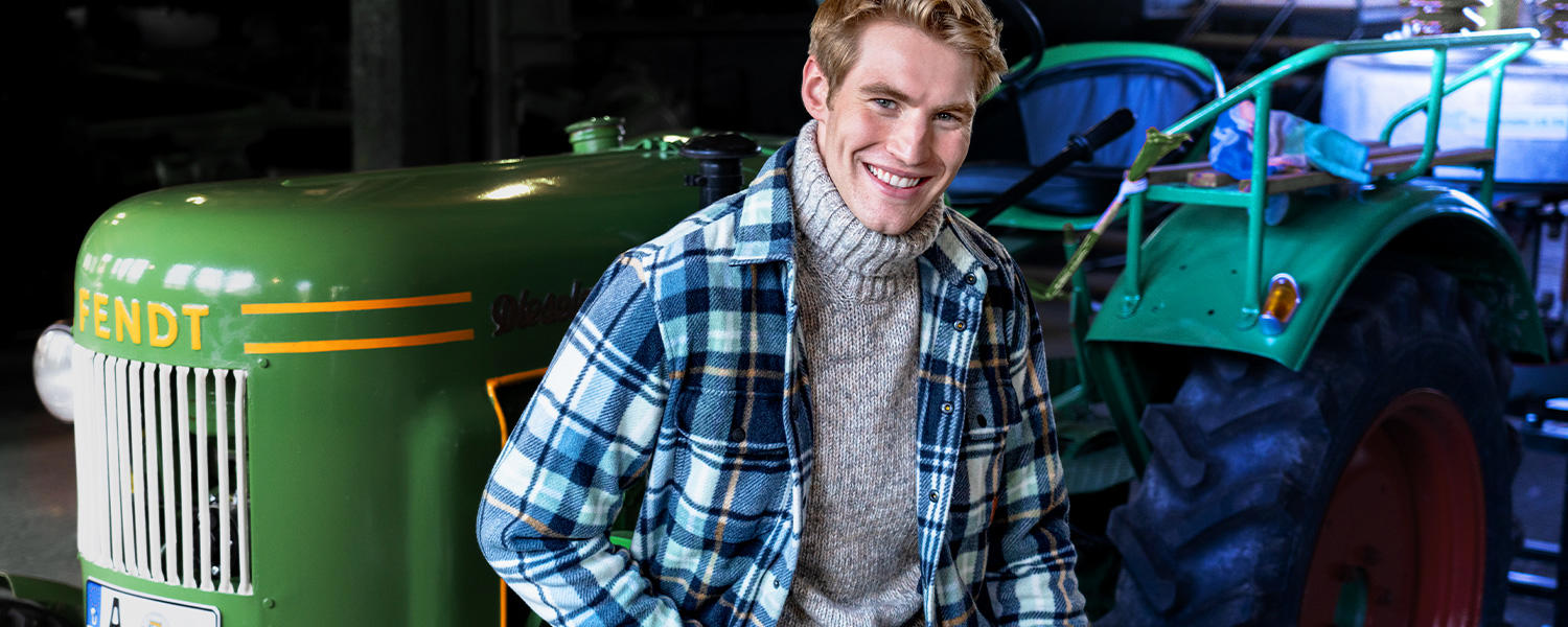 Ein junger Mann steht vor einem grünen Fendt-Traktor. Er trägt einen grauen Grobstrick-Rollkragenpullover und ein blau-beiges kariertes Overshirt, was ländliche Gemütlichkeit ausstrahlt.