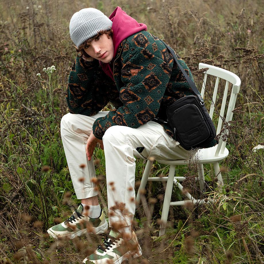 Ein junger Mann sitzt auf einem weißen Holzstuhl auf einer Wiese. Er trägt eine hellgraue Mütze, einen roten Hoodie mit einer dunkelgrünen Teddyfell-Jacke darüber und eine beige Chino.
