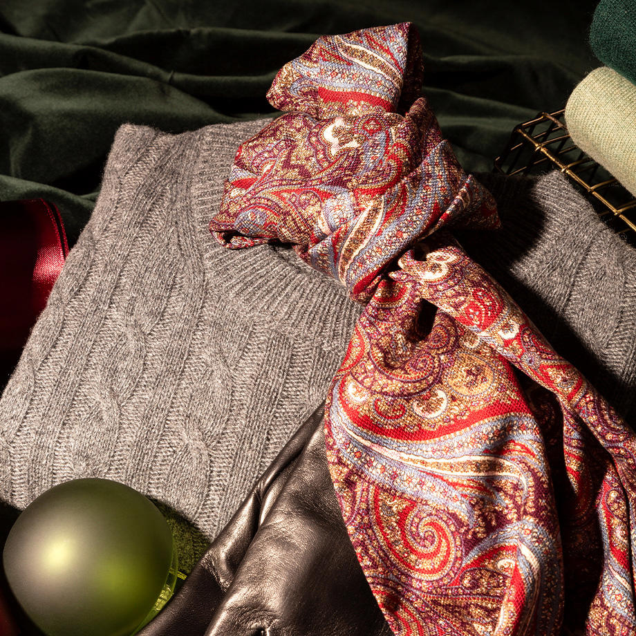 Ein Arrangement aus eleganter Herrenmode mit einem grauen Strickpullover, einem luxuriösen roten Paisley-Schal und schwarzen Lederhandschuhen. Im Hintergrund sorgen grüne Stoffe und eine matte, grüne Kugel für eine festliche und warme Atmosphäre.