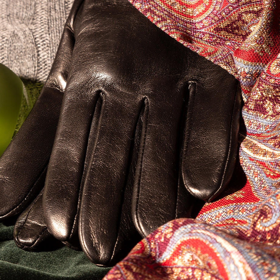 Schwarze Lederhandschuhe auf einem bunten Paisley-Stoff, kombiniert mit einem grauen Strickpullover und einem grünen Hintergrund für einen luxuriösen und warmen Winterlook.