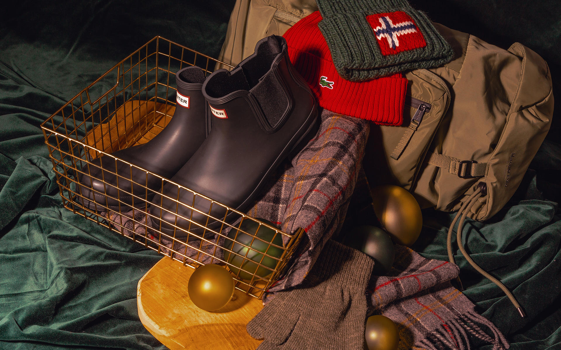Auf dem Bild sieht man auf einer Samtdecke in Grün einen goldenen Korb mit Gummistiefel und einem karierten Schal. Außerdem sind auf dem Bild zwei Mützen, Handschuhe, eine Reisetasche und Weihnachtskugeln zu sehen.