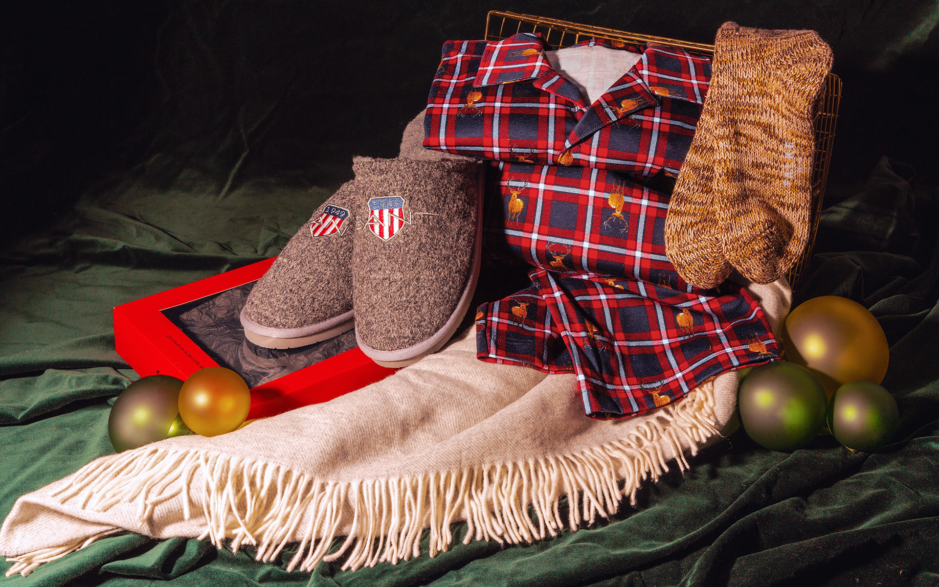 Zu sehen sind ein Pyjama, kuschelige Socken, ein heller Schal und Pantoffeln, umgeben von Weihnachtskugeln