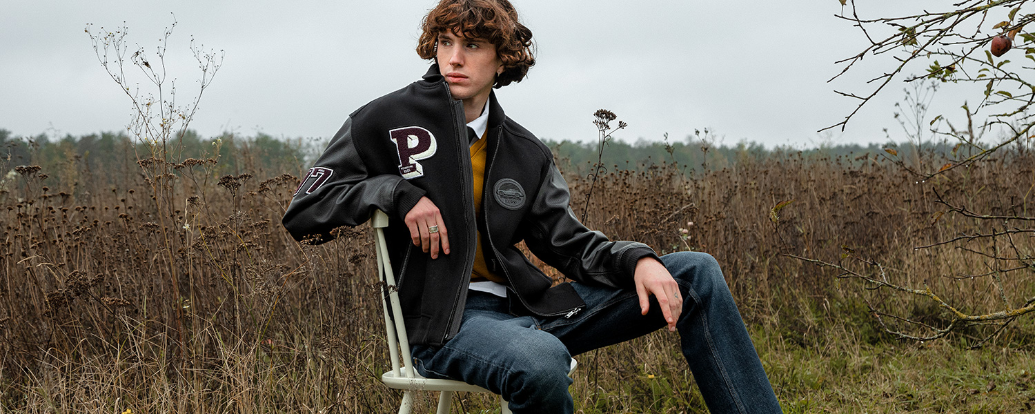 ein junger Mann in Collegejacke sitzt auf einem weißen Stuhl vor einem Feld
