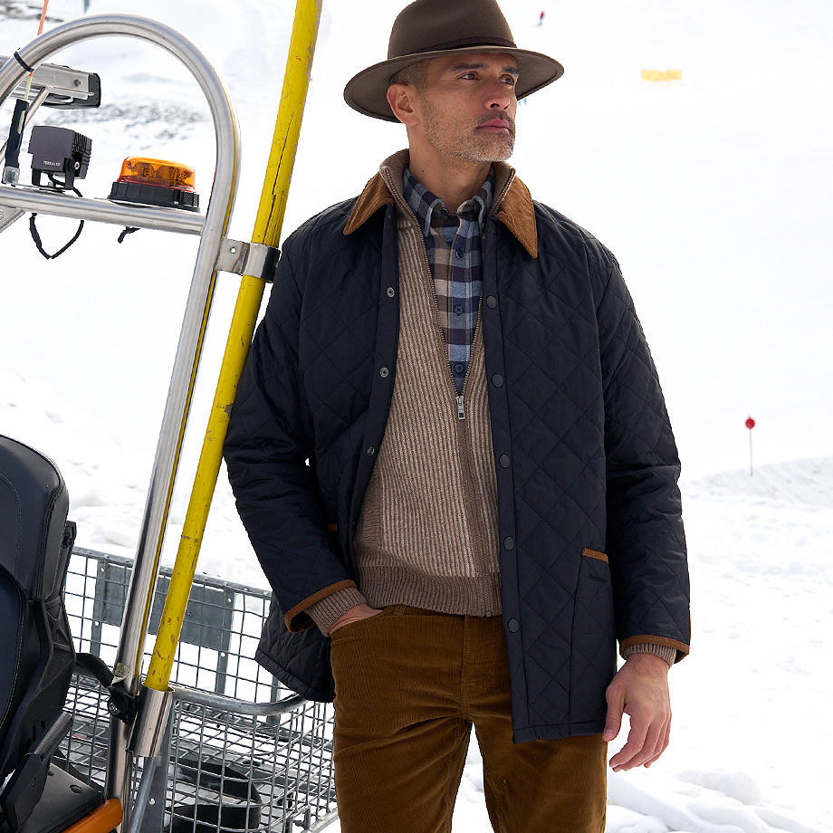 Ein Mann steht im Schnee und hat die rechte Hand in der Hosentasche seiner Cordhose. Er trägt einen Hut, ein kariertes Hemd, eine Strickjacke und eine Steppjacke. 