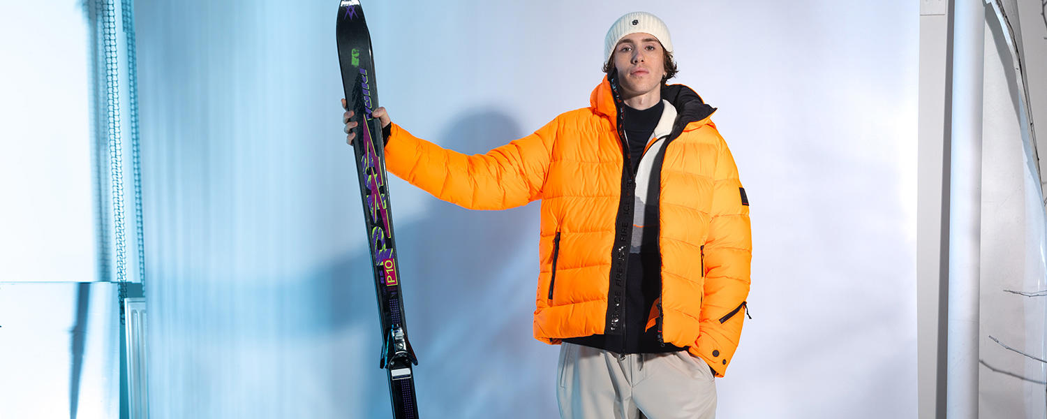 Mann in neon-orangener Winterjacke, weißer Mütze und grauer Hose steht vor einem neutralen Hintergrund mit eisblauem Licht. In der einen Hand hält er ein Paar Ski.