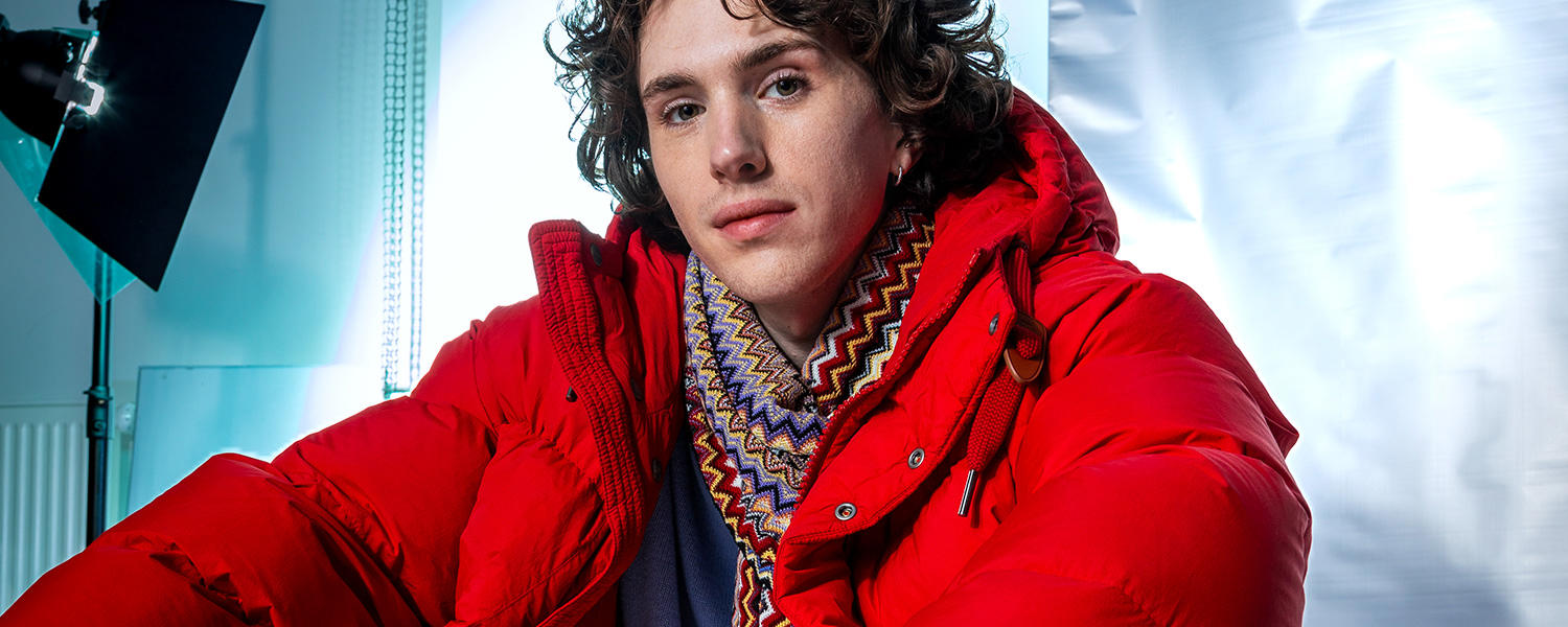 ein junger Mann trägt eine knallrote Winterjacke und einen bunten Schal. Er sitzt in einem Fotostudio