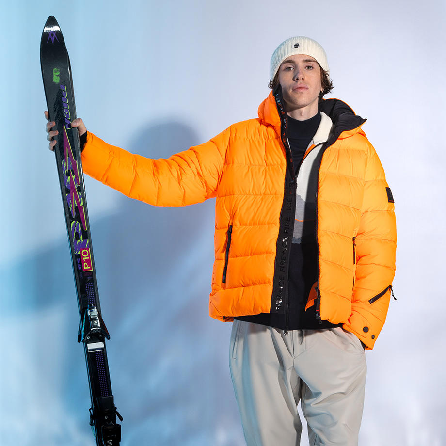 Ein junger Mann steht vor einem eisblauen Hintergrund. In der einen Hand hält er ein Paar Ski. Er trägt eine neonorange Winterjacke, eine graue Hose und eine weiße Beanie.