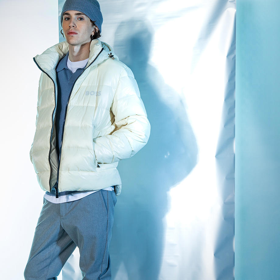 Ein junger Mann steht vor einem eisblauen Hintergrund. Er trägt eine weiße Winterjacke, eine graue Chino und eine graue Beanie.