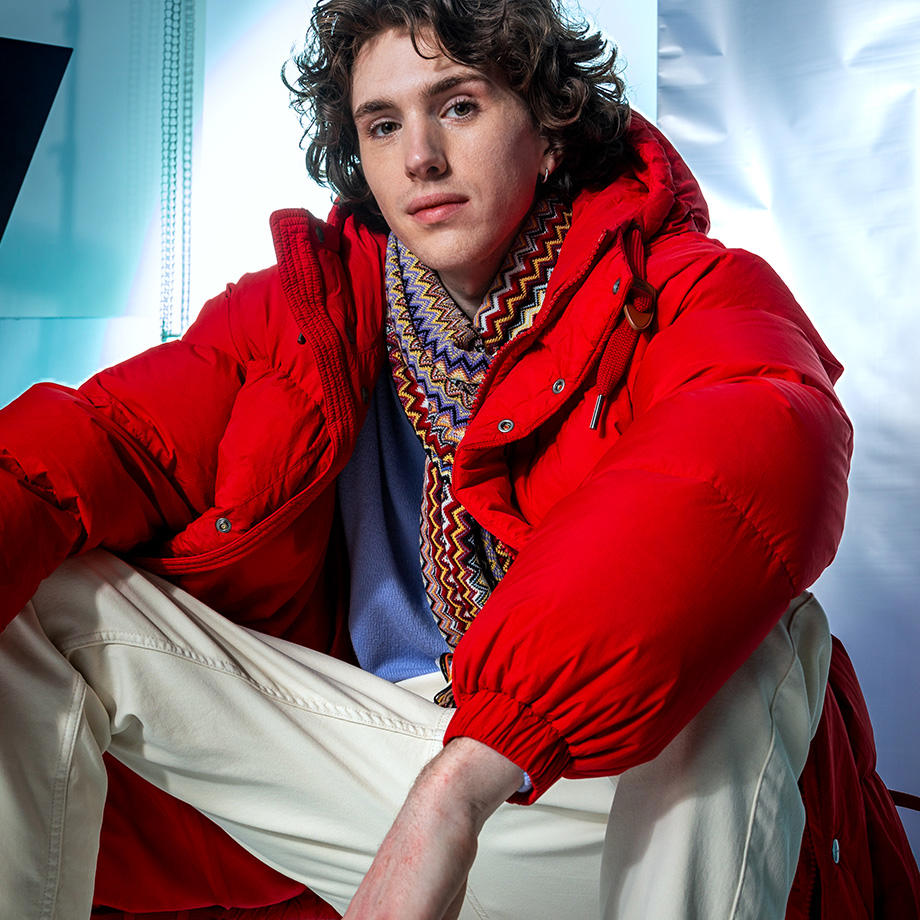 Ein junger Mann sitzt in einem Studio. Er trägt eine knallrote Pufferjacket und einen bunten Schal