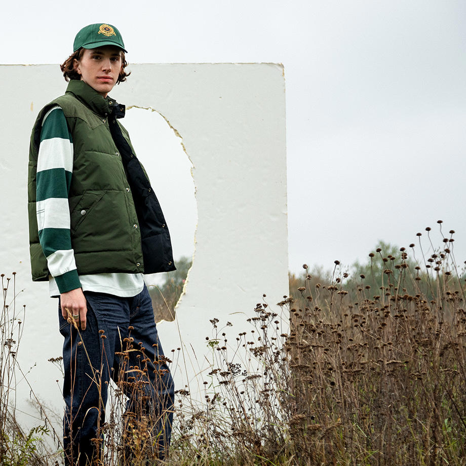 Ein junges männliches Model auf einem abgeernteten Feld mit einer weißen Pappe im Hintergund. Er trägt ein gestreiftes Rugby-Shirt mit einer olivfarbenen Weste darüber.