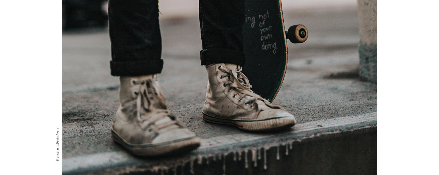 Auf dem Bild sieht man dreckige Sneakers an den Füßen einer Person mit einem Skateboard in der Hand.