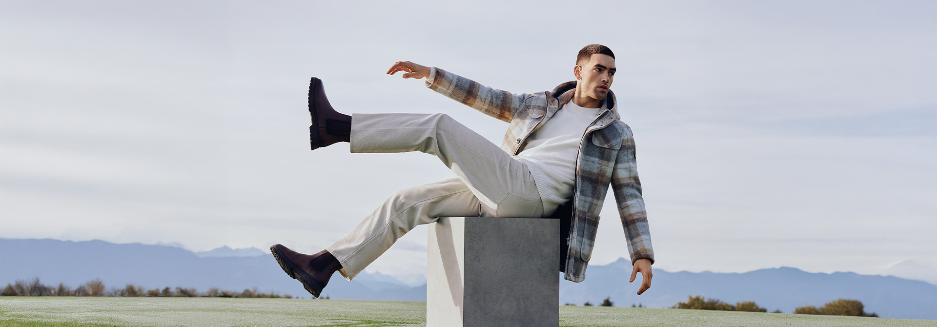 Ein Mann trägt einen karierten Wollmantel. Er sitzt auf einem Betonblock auf einer Wiese. Er hat die Arme und beide Beine in der Luft, als würde er nach hinten kippen.