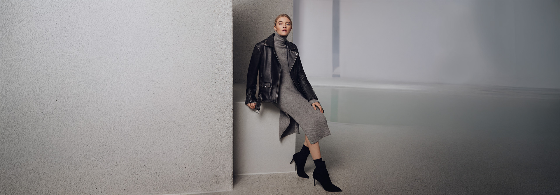 Eine Dame trägt ein graues Kleid und eine schwarze Lederjacke. Sie lehnt an einer grauen Betonwand.