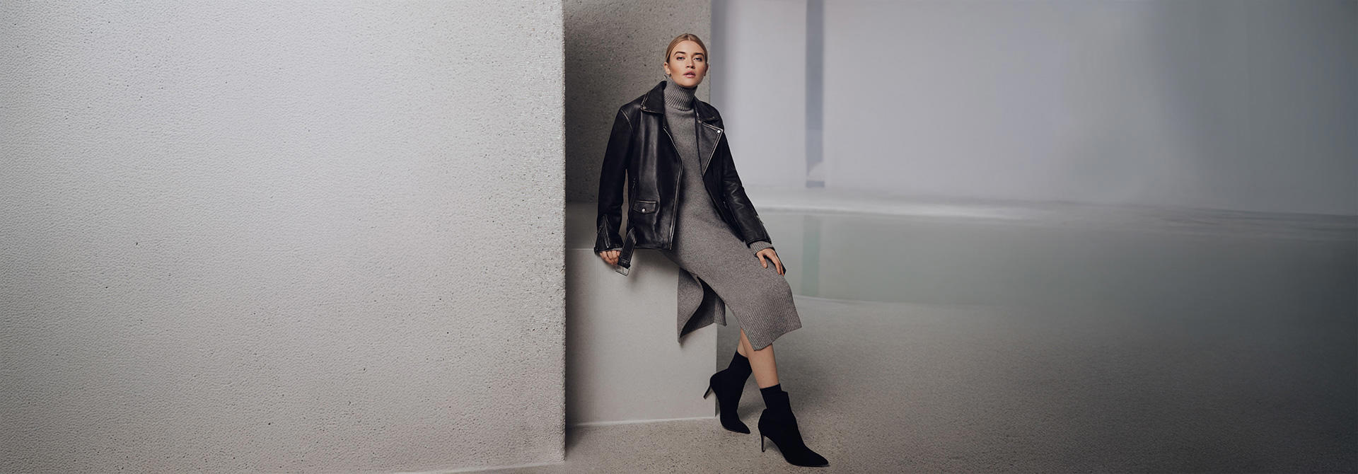 Eine Dame trägt ein graues Kleid und eine schwarze Lederjacke. Sie lehnt an einer grauen Betonwand.