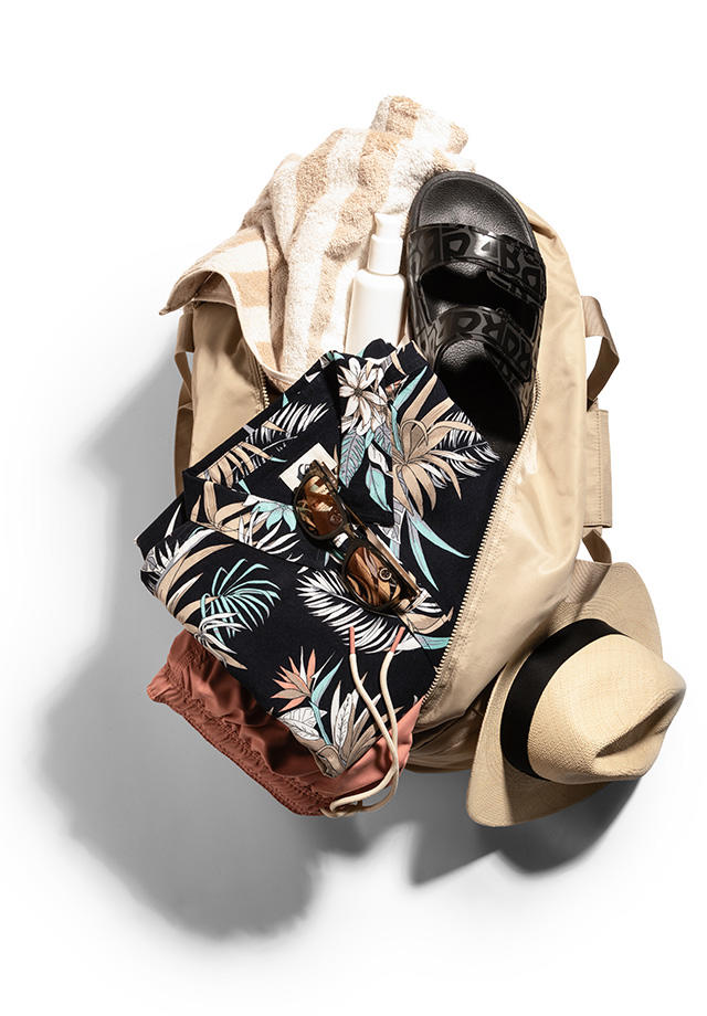 Eine halb geöffnete Reisetasche. In ihr sind ein florales Hemd, eine Badehose, ein Strandtuch, eine Sandale und eine Sonnenbrille zu sehen. Am Rand lehnt ein Sonnenhut.