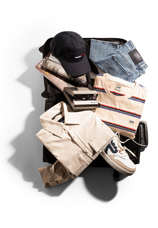 Ein geöffneter Trolli. In ihm liegen eine Jeans, eine Cap, ein gestreiftes T-Shirt, ein leichtes Overshirt, ein Sneaker, ein paar Magazine und eine analoge Kamera