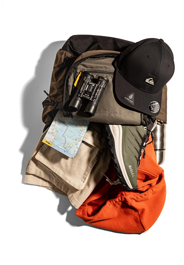 Ein geöffneter Rucksack. In ihm sind ein Wanderschuh, ein oranger Hoodie, eine Cargoshorts, eine Landkarte und ein Fernglas zu sehen. Auf ihr drauf liegt eine Cap.