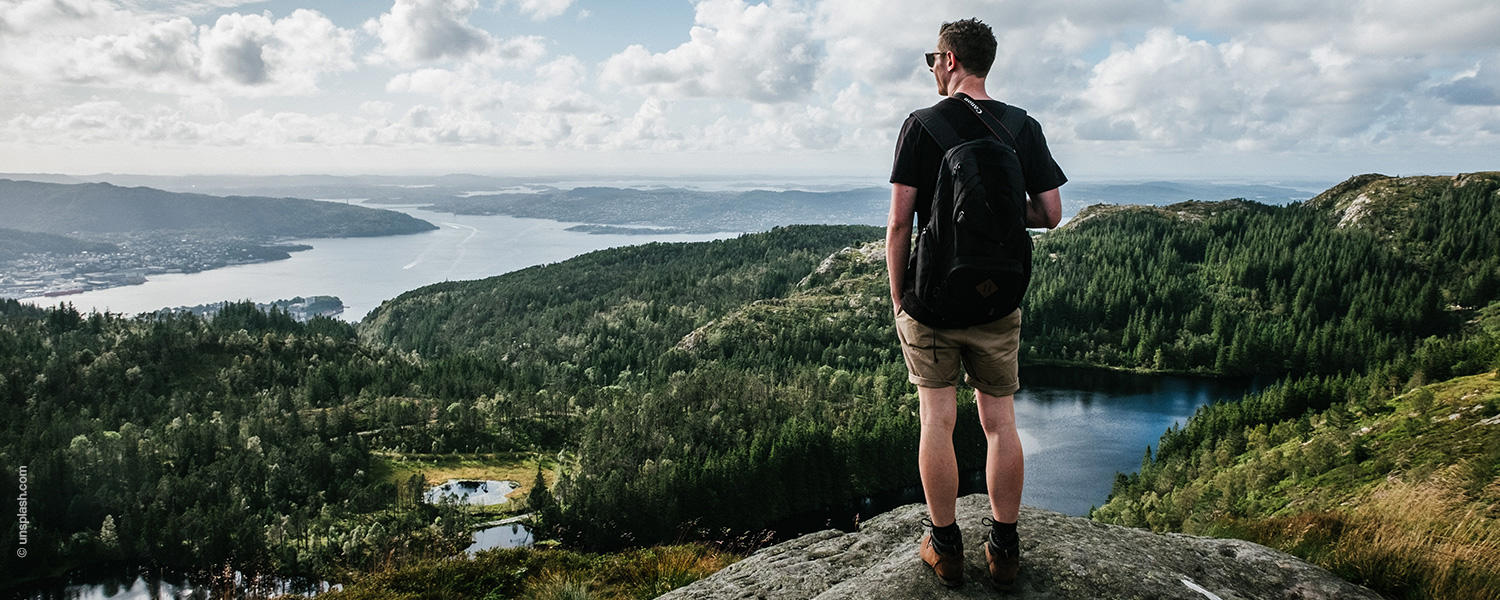 Mann steht auf einem Felsen draußen in der Natur. Er trägt eine kurze Hose, T-Shirt und hat einen Rucksack.