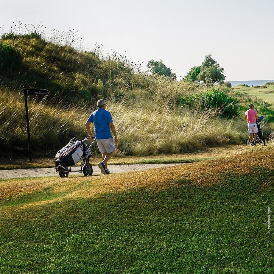 Das Bild zeigt drei Männer, die mit Golfwagen einen Pfad entlanglaufen, umgeben von hohen Gräsern und einer weiten Golfplatzlandschaft.