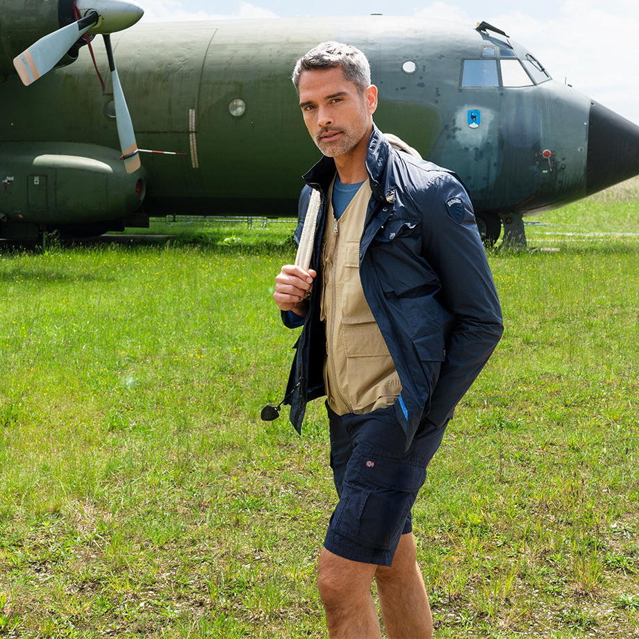 Männliches Model auf einer Wiese mit einer Cargo-Short, Weste und Jacke. 
