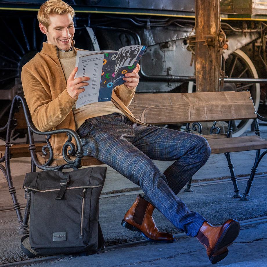 Ein Mann sitzt auf einer Holzbank, trägt eine blau karierte Hose, braune Chelsea Boots, einen beigefarbenen Pullover und dazu passend eine beige Jacke. 