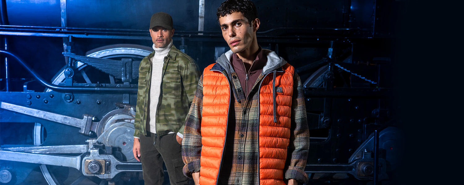 Der Mann im Vordergrund trägt eine orange Steppweste über einem karierten Overshirt und einem Troyer, kombiniert mit hellgrauen Jeans; der Mann im Hintergrund trägt eine olivgrüne Cargohose, einen camo-grünen Blouson und einen cremefarbenen Rollkragenpullover, abgerundet durch eine grüne Kappe.