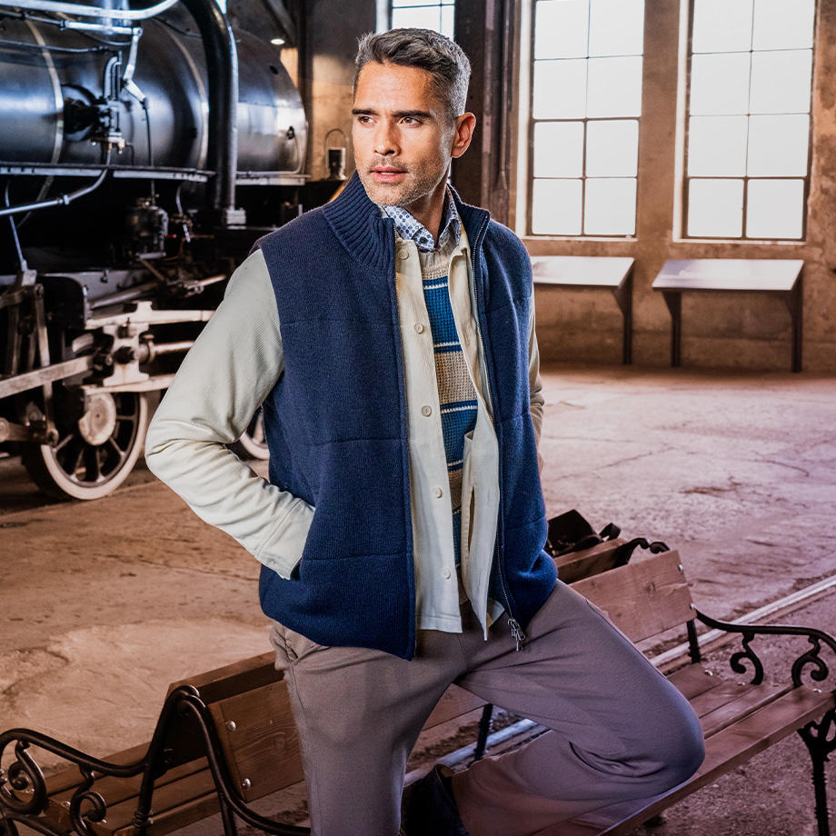Ein Mann im Lagenlook mit einer blauen Weste, einem beigen Overshirt und einem beige-blau gestreiften Pullover, ergänzt durch eine graue Hose