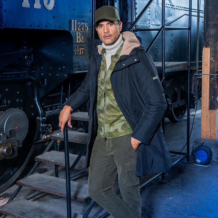  Ein Mann trägt einen dunkelblauen Parka über einem Camouflage-Overshirt und einem grauen Rollkragenpullover, ergänzt durch olivfarbene Cargohosen