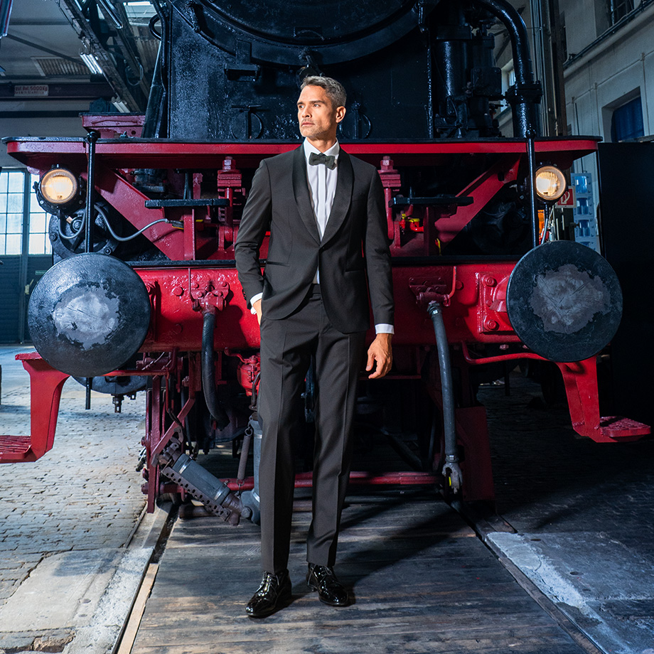 Ein Mann mit einem Smoking, Krawatte und Fliege. Die rechte Hand hat er lässig in die Hosentasche gesteckt. 