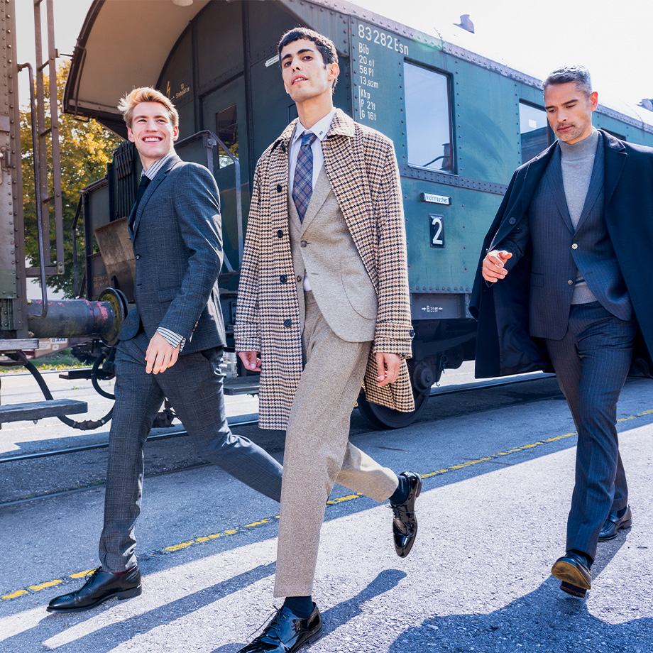 Drei Männer laufen an einem Bahn-Abteil vorbei.Der mittlere Mann trägt eine karierten Mantel mit hellem Anzug, der rechte Mann trägt einen Karo-Anzug und der linke Mann eine Mantel-Anzug-Kombination. 