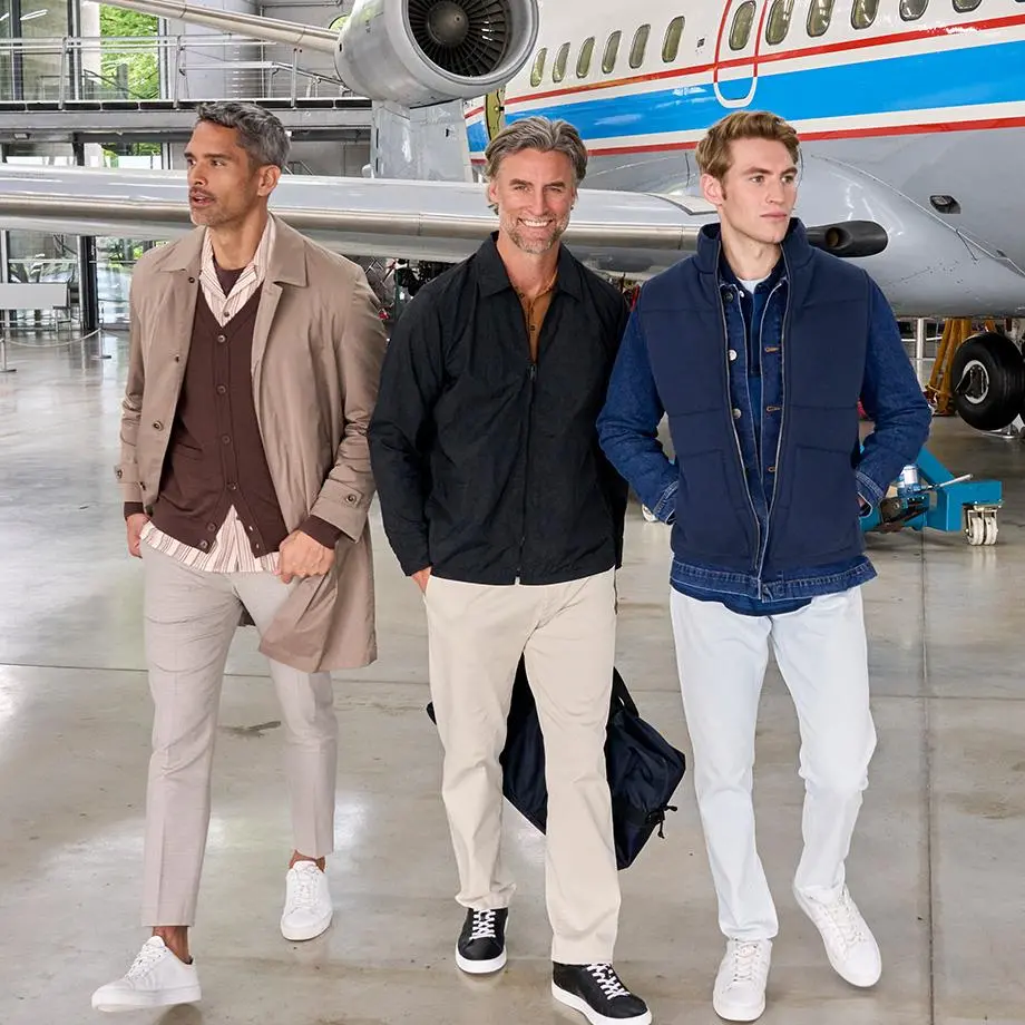 Das Bild zeigt drei Männer in Basic-Outfits: Der erste trägt helle Jeans, eine blaue Jeansjacke und eine blaue Weste, der zweite eine schwarze Jacke, beige Hose und schwarze Sneaker, und der dritte einen Trenchcoat mit brauner Strickjacke, gestreiftem Hemd, heller Hose und weißen Sneakern.