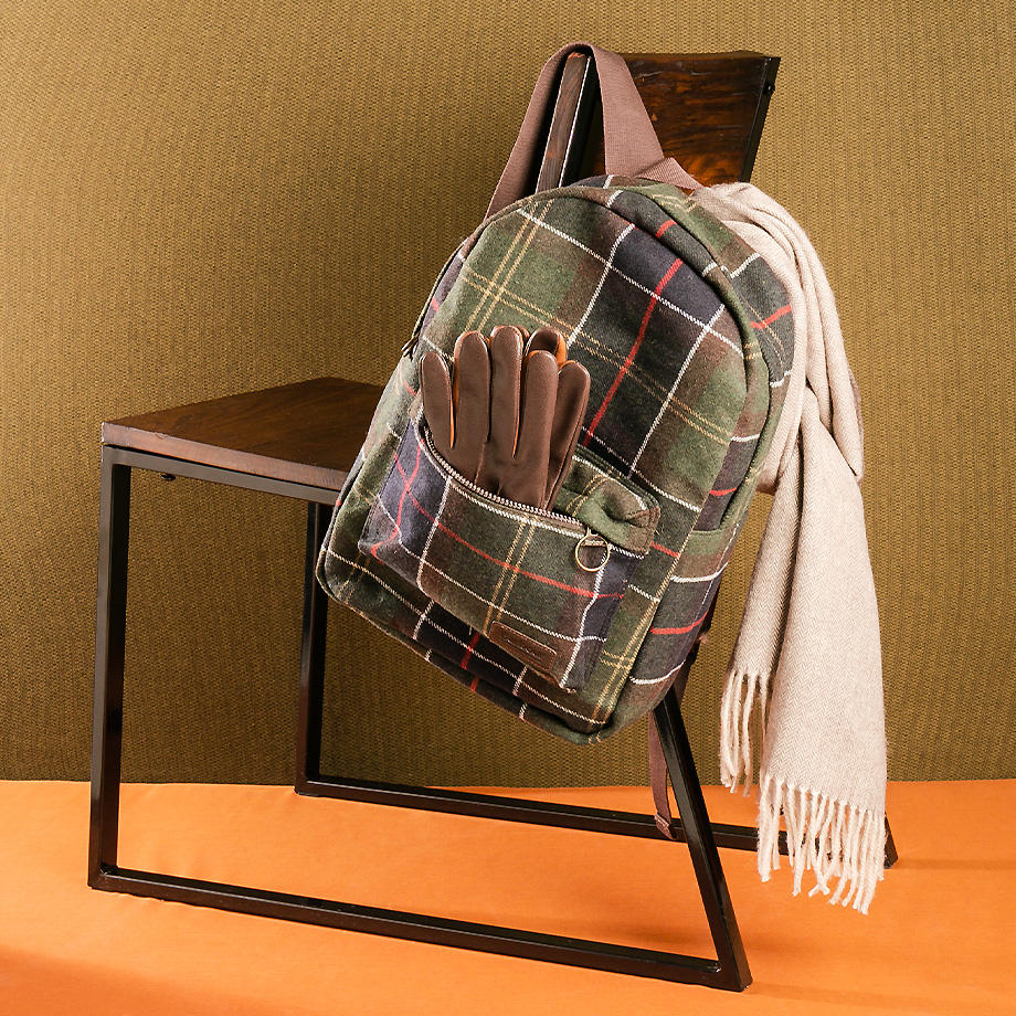 Das Bild zeigt einen karierten Rucksack mit Lederdetails, braunen Handschuhen und einem beigen Schal die über eine Stuhllehne drapiert sind. 