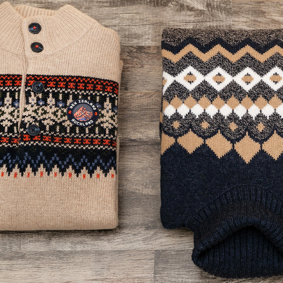 Zwei gestrickte Pullover mit nordischem Muster, einer in Beige mit dunkelblauen und weißen Akzenten, der andere in Dunkelblau mit beige-weißen Mustern, auf einer rustikalen Holzoberfläche.