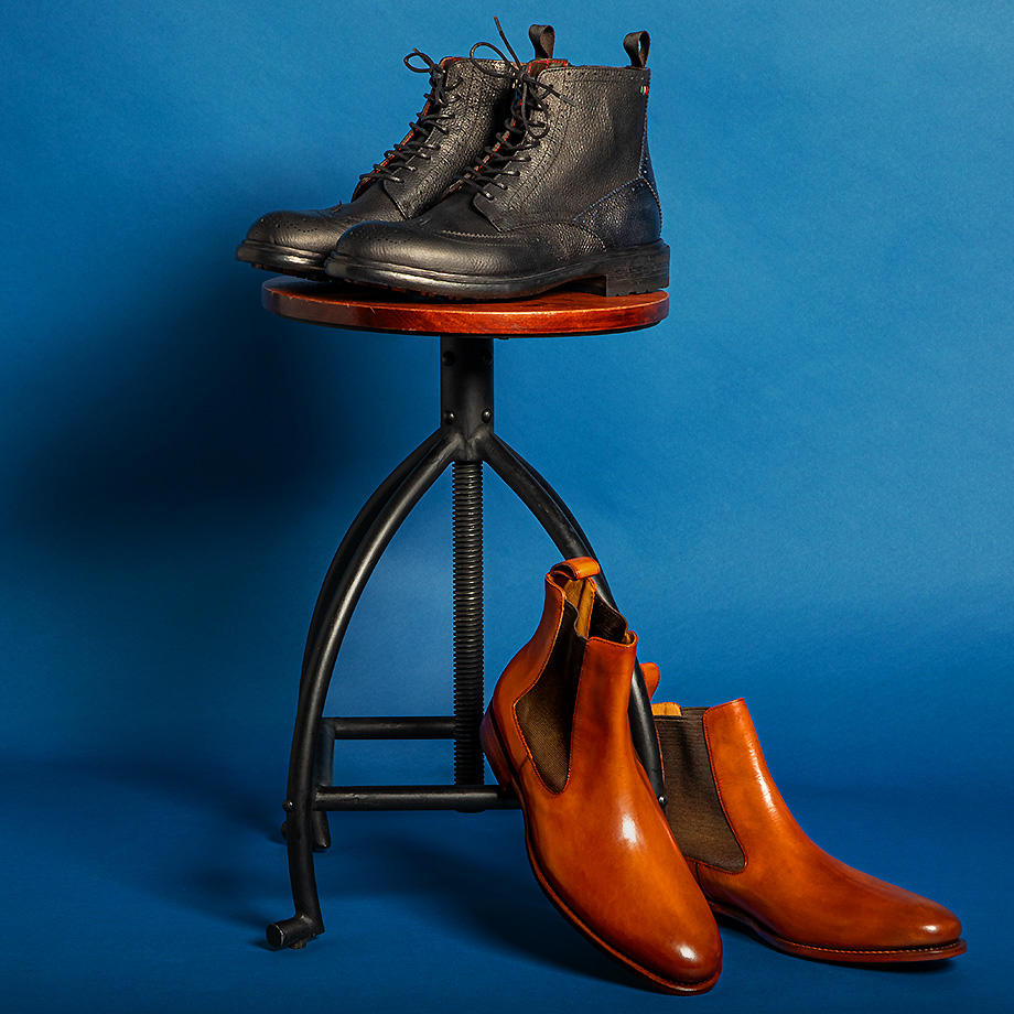 Das Bild zeigt schwarze Lederstiefel vor blauem Hintergrund, die auf einem runden Hocker stehen. Cognacfarbene Chelsea-Boots stehen am Boden an den Hocker gelehnt. 