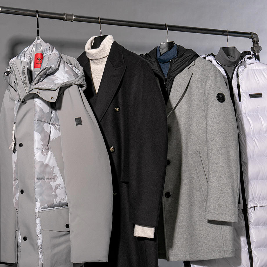 Eine vielseitige Auswahl an Winterjacken und Mänteln – darunter ein eleganter schwarzer Wollmantel, ein grauer Wollmantel, ein grauer Parka und eine gesteppte Daunenjacke – stilvoll an einer Kleiderstange präsentiert.