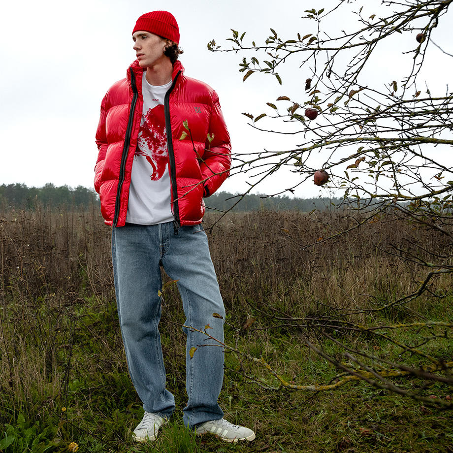 Ein junger Mann steht auf einem Feld im Herbst. Er trägt eine knallrote Pufferjacket mit passender Mütze, darunter ein T-Shirt mit rotem Print und eine weite blaue Jeans.