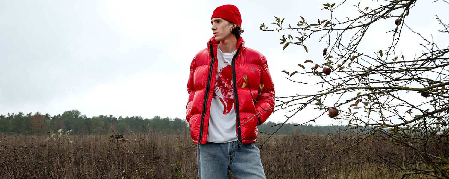Ein junger Mann steht auf einem Feld im Herbst. Er trägt eine knallrote Pufferjacket mit passender Mütze und darunter ein T-Shirt mit rotem Print.