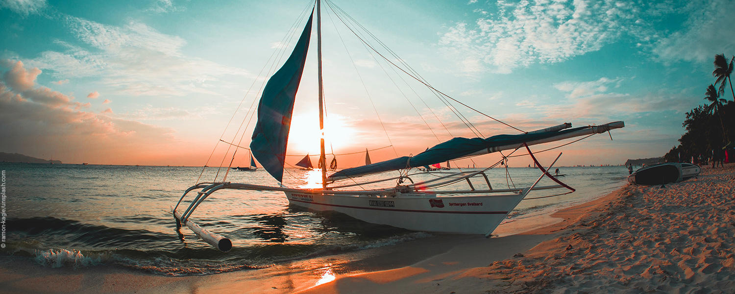 Ein Segelboot liegt am Strand und dahinter geht die Sonne unter. 