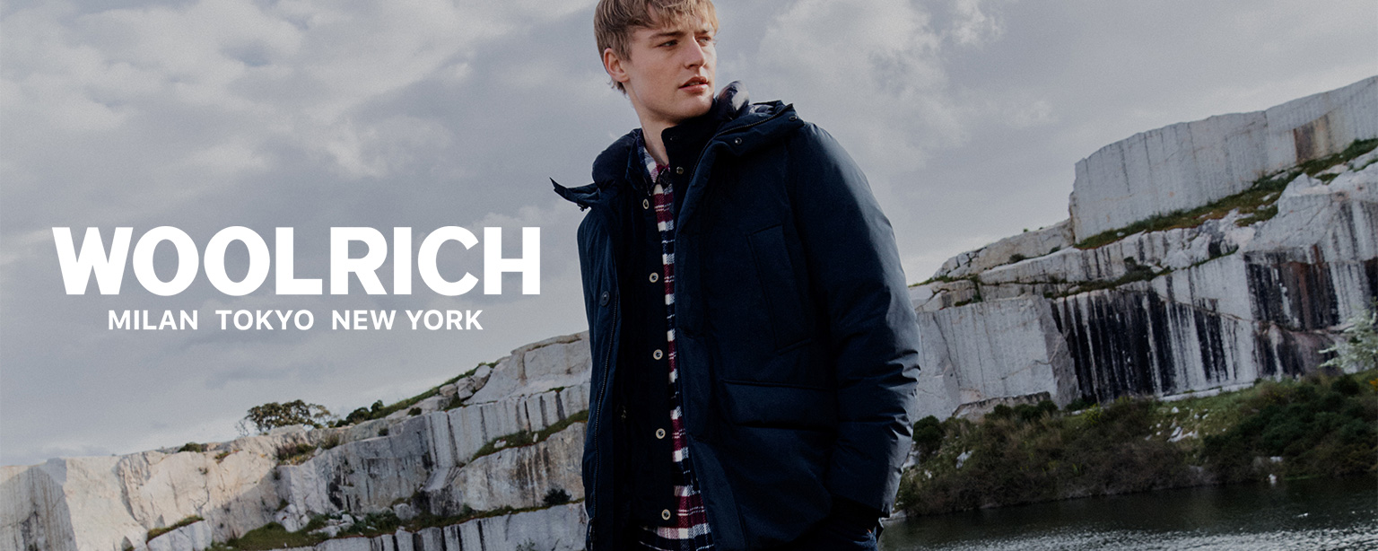 Woolrich Herrenmode – Outdoor-Look mit warmer Winterjacke, kariertem Hemd und Boots in natürlicher Felslandschaft am Wasser.