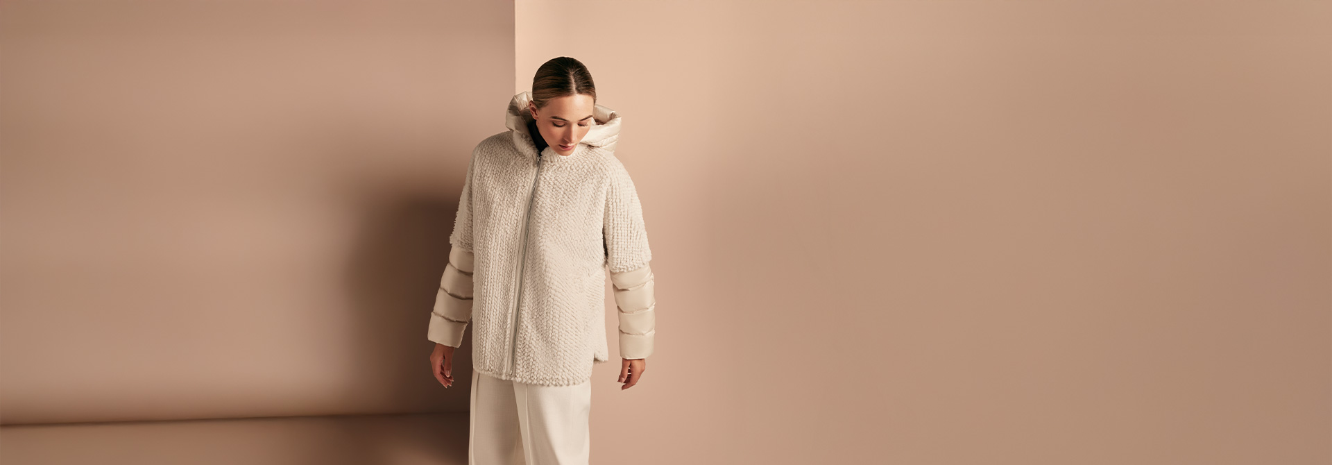 Eine Dame trägt eine Winterjacke aus Kunstfell mit abgesetzten Ärmeln aus Stepp vor beigem Hintergrund.