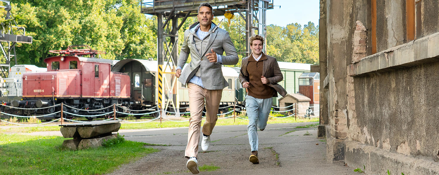 Zwei Männer in stilvoller Freizeitkleidung laufen lachend durch ein Industrieareal. Der Mann vorn trägt ein hellgraues Sakko, ein hellblaues Hemd und beige Chinos, der Mann dahinter ein braunes Jackett mit weißem Hemd und hellblauer Hose. Im Hintergrund sind alte Eisenbahnwaggons und grüne Bäume zu sehen.
