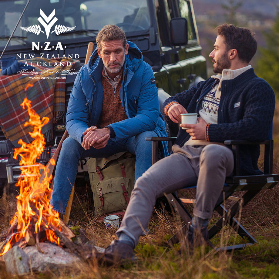 Männer im Gespräch, tragen blaue Daunenjacke und Norweger-Pullover, sitzen am Lagerfeuer.