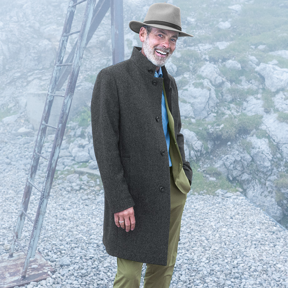 Älterer Mann mit grauem Hut und dunklem Mantel steht lächelnd in einer nebligen Berglandschaft – stilvolle Herbst- und Wintermode für Herren.